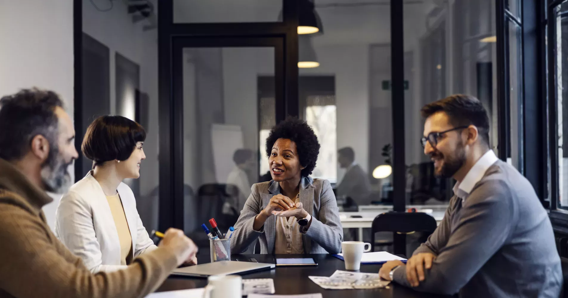 Group of professionals collaborating around a wooden conference table in a modern office with large windows, natural light, and city views, creating a bright and productive team environment.