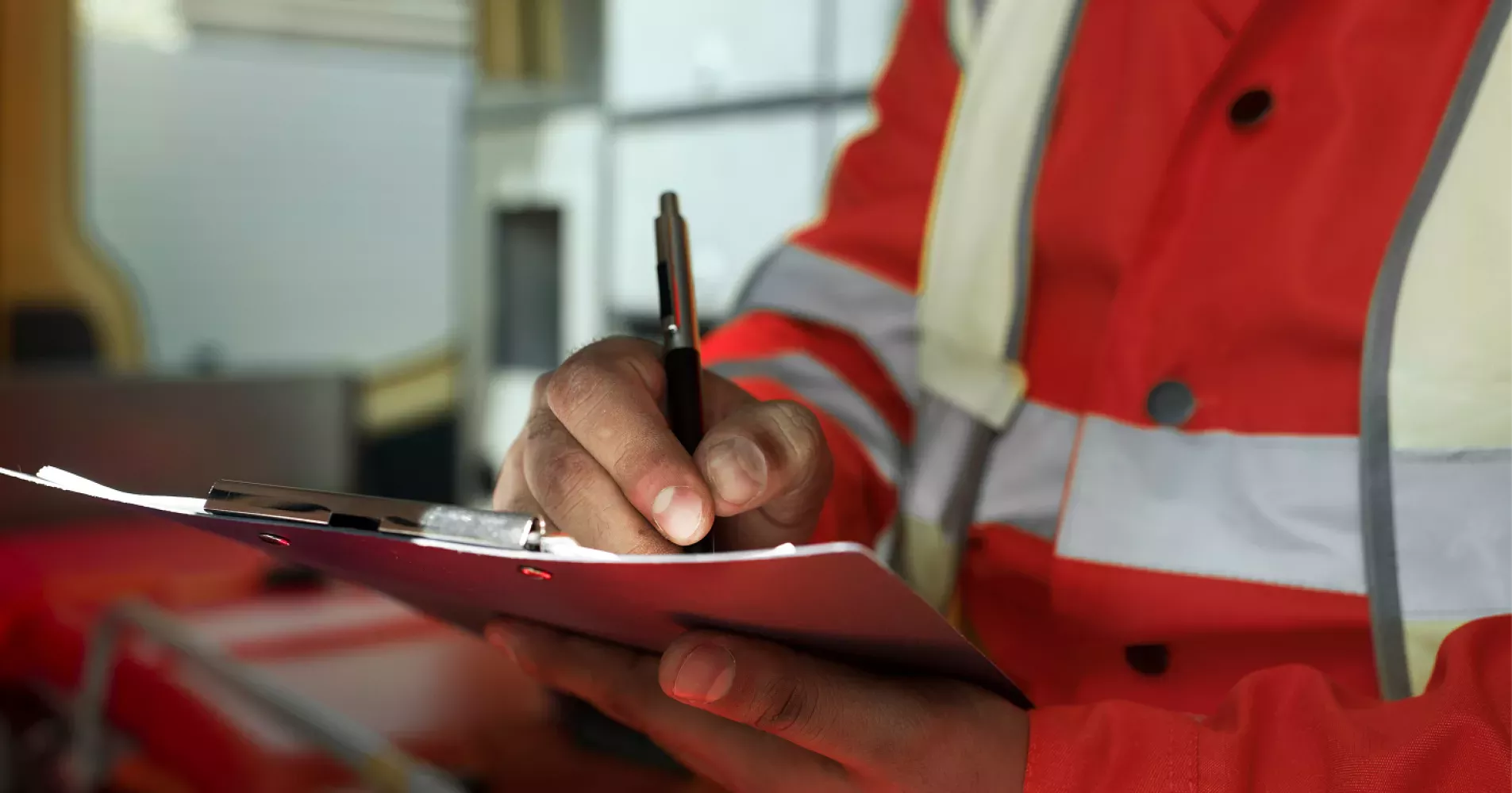 Close-up of a worker in a red and reflective safety jacket writing on a clipboard, possibly conducting an inspection or taking notes inside or near an emergency response vehicle.