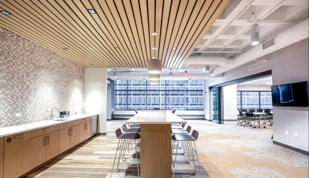 Stylish communal break area at 1818 Market featuring a long high-top table with modern barstools, wood-paneled ceiling with pendant lighting, a sleek kitchenette with herringbone tile backsplash, and an adjacent flexible conference room with glass walls and city views.
