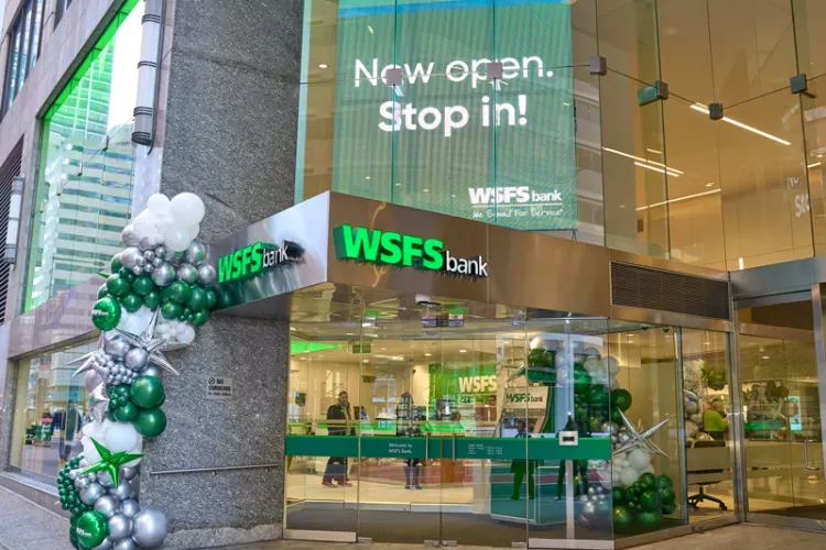 Street-level entrance of the WSFS Bank branch at 1818 Market, featuring a vibrant grand opening display with green and silver balloons, a modern glass storefront, and a large digital sign reading “Now open. Stop in!” inviting customers inside.